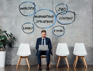 smiling businessman sitting on chair and using laptop in waiting hall with marketing strategy