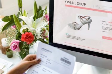 Piece of paper in womans hand while online shopping for stiletto style shoes beside vase of flowers