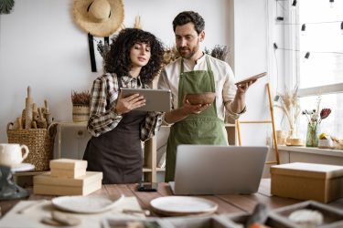 Bearded mature man counting goods while young curly woman using tablet. Two Caucasian workers in uniform doing inventory together at decor shop.