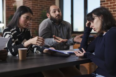 Tired recruiter listening to irritated job applicants complaining about absurd salary offer while sitting at desk in office space. Exhausted businesswoman reviewing candidates CV resumes.