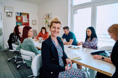A diverse team of business professionals engaged in a discussion about projects in a modern glass walled office.